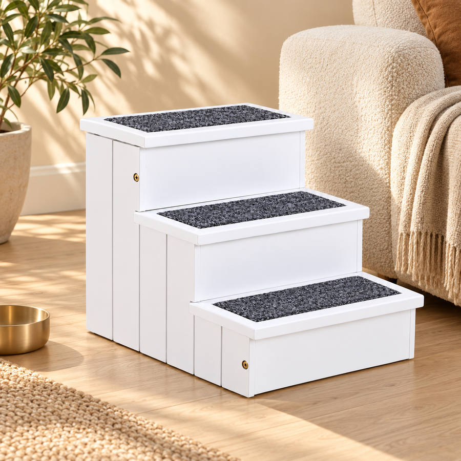 White three-step wooden pet stairs with dark carpet treads beside a cream armchair on a wood floor