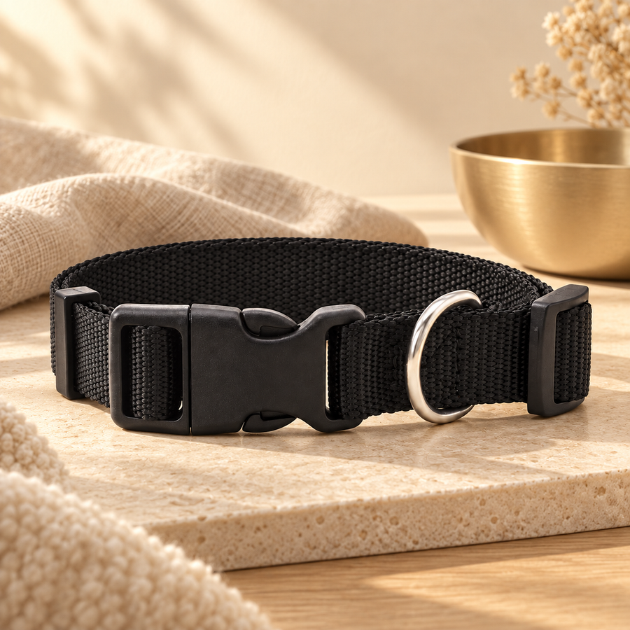 Black nylon dog collar with a buckle resting on a stone tray beside a knit throw and a brass bowl