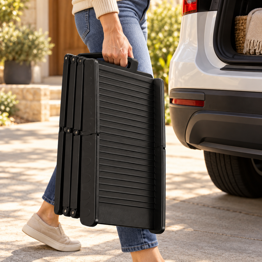 Person carrying a folded black tri-fold dog ramp by its handle next to a white SUV