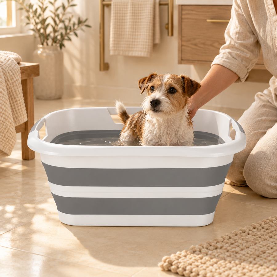 Small terrier standing in a gray and white bathing tub while an adult kneels beside it on a bathroom floor.