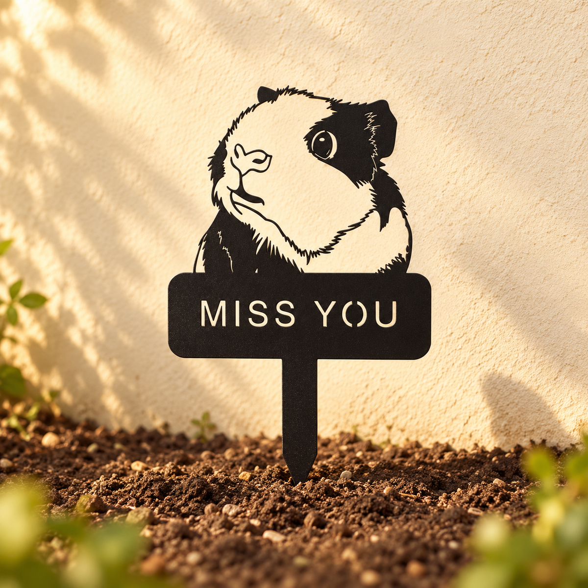 Guinea pig memorial stake with a MISS YOU inscription standing in soil against a warm cream wall