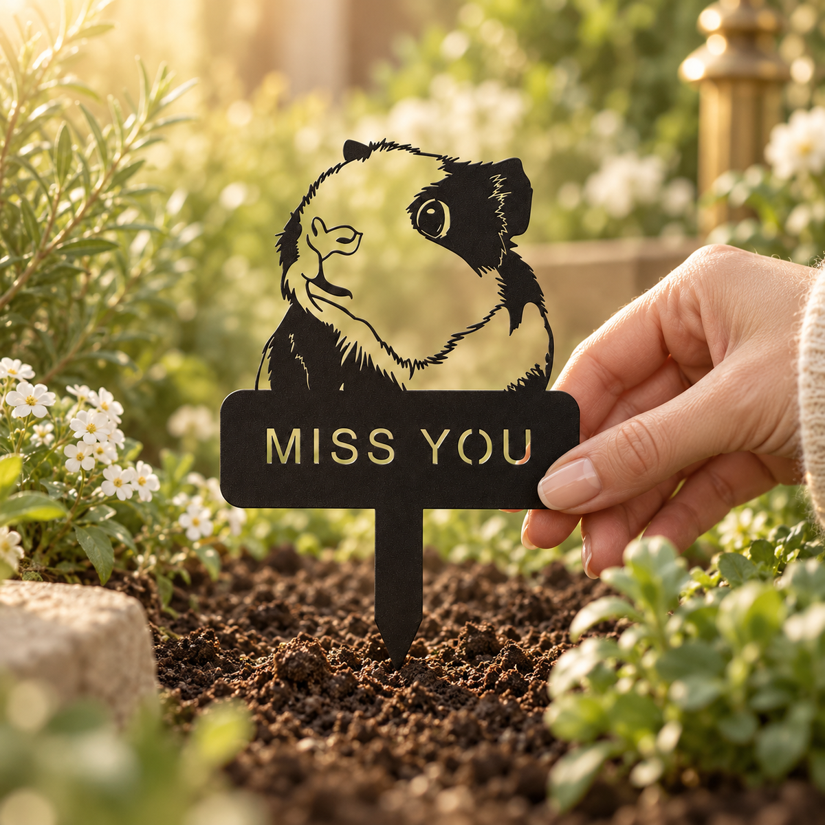 Hand holding the edge of a black MISS YOU memorial plaque above its pointed stem in garden soil